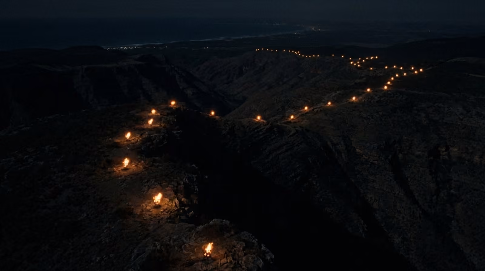 Aerial night view: chains of beacon fires strung along mountains toward a coastline.